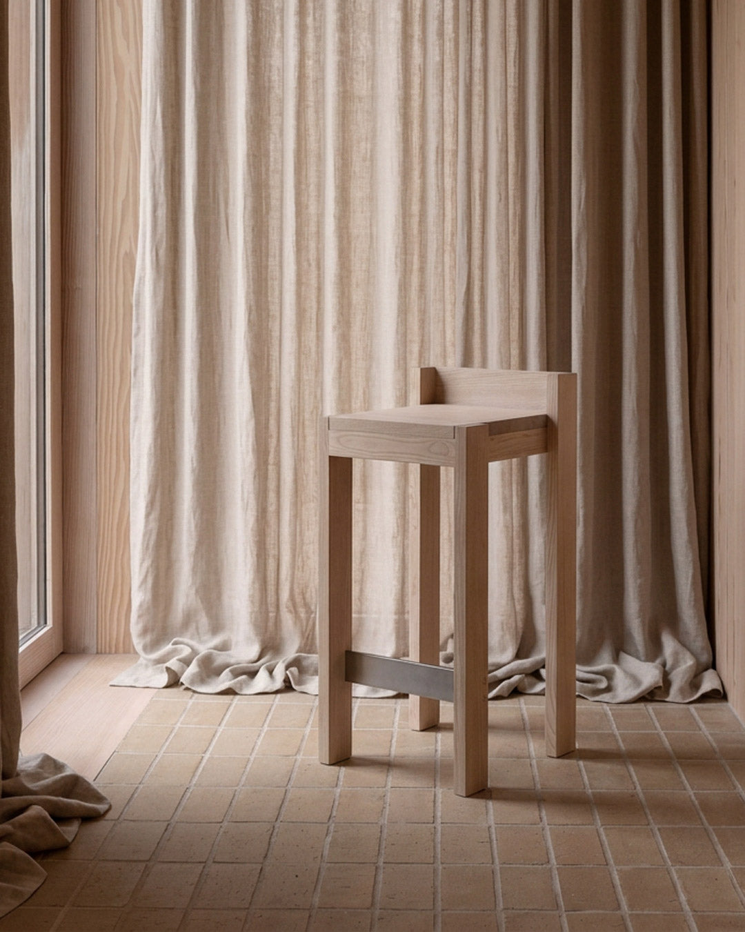 A minimalist light wood stool with a square backrest and a metal support bar, set in a warm, neutral-toned room with beige curtains and tiled flooring.