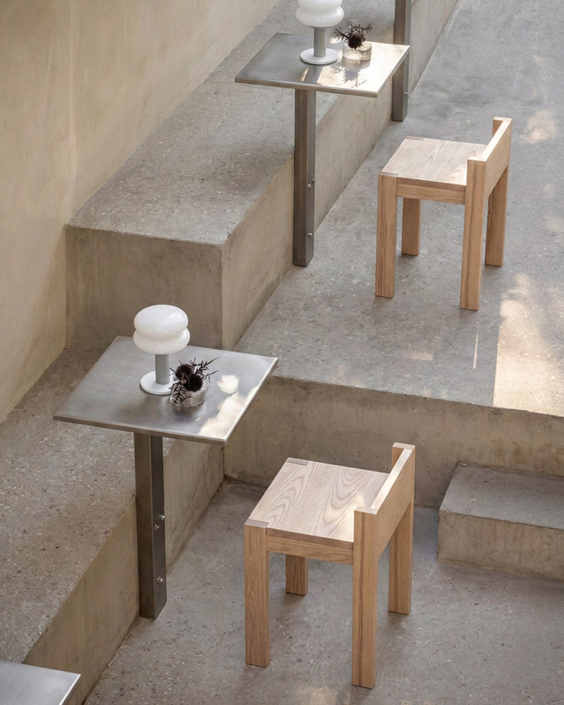 Minimalist outdoor seating featuring light natural wood chairs and square stainless steel tables arranged on concrete steps.