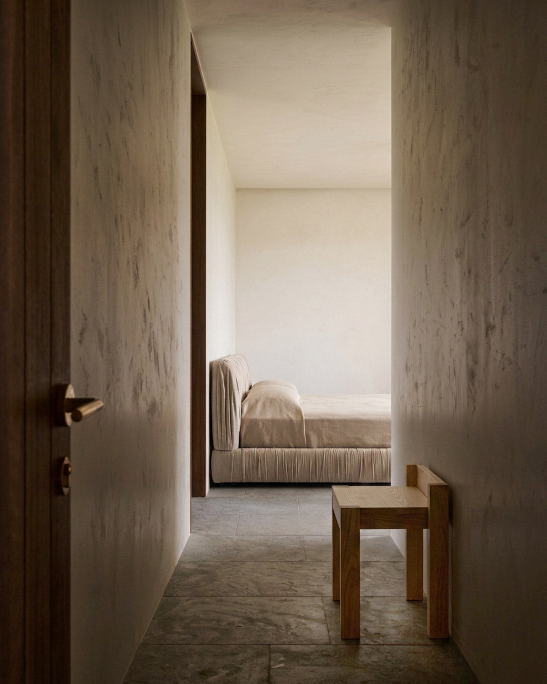 A minimalist bedroom scene viewed from a hallway, featuring a low wooden stool in the foreground, textured plaster walls, and a bed with neutral beige bedding in the background.