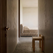 A minimalist bedroom scene viewed from a hallway, featuring a low wooden stool in the foreground, textured plaster walls, and a bed with neutral beige bedding in the background.
