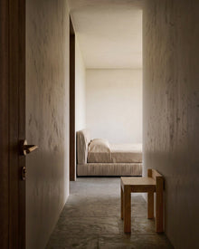 A minimalist bedroom scene viewed from a hallway, featuring a low wooden stool in the foreground, textured plaster walls, and a bed with neutral beige bedding in the background.