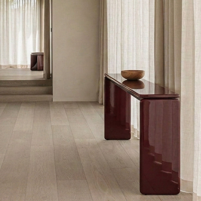 A modern console table with a glossy dark burgundy finish and a rectangular wood bowl on top, set in a minimalist room with light wood flooring and sheer curtains.