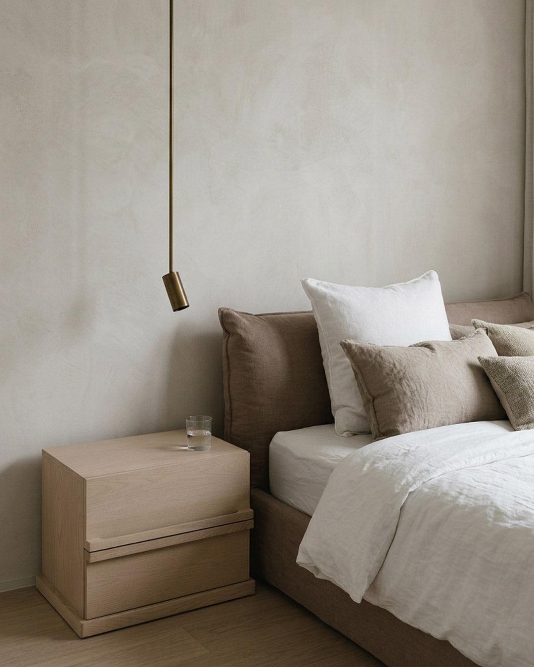 A minimalist bedroom scene featuring a light wood two-drawer nightstand next to a bed with white and beige linens, set against a textured beige plaster wall with a hanging brass pendant light.