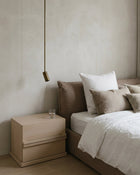 A minimalist bedroom scene featuring a light wood two-drawer nightstand next to a bed with white and beige linens, set against a textured beige plaster wall with a hanging brass pendant light.