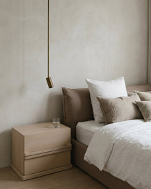 A minimalist bedroom scene featuring a light wood two-drawer nightstand next to a bed with white and beige linens, set against a textured beige plaster wall with a hanging brass pendant light.