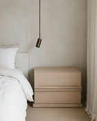 A light natural wood two-drawer nightstand in a minimalist bedroom setting next to a bed with white bedding and a hanging brass pendant light.