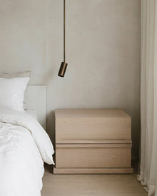 A light natural wood two-drawer nightstand in a minimalist bedroom setting next to a bed with white bedding and a hanging brass pendant light.