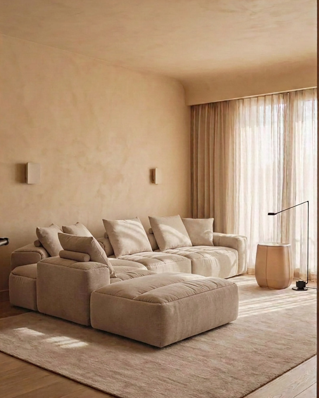 A modern modular sectional sofa in a warm beige fabric, styled in a minimalist living room with a matching ottoman, textured beige walls, sheer curtains, and a large area rug.