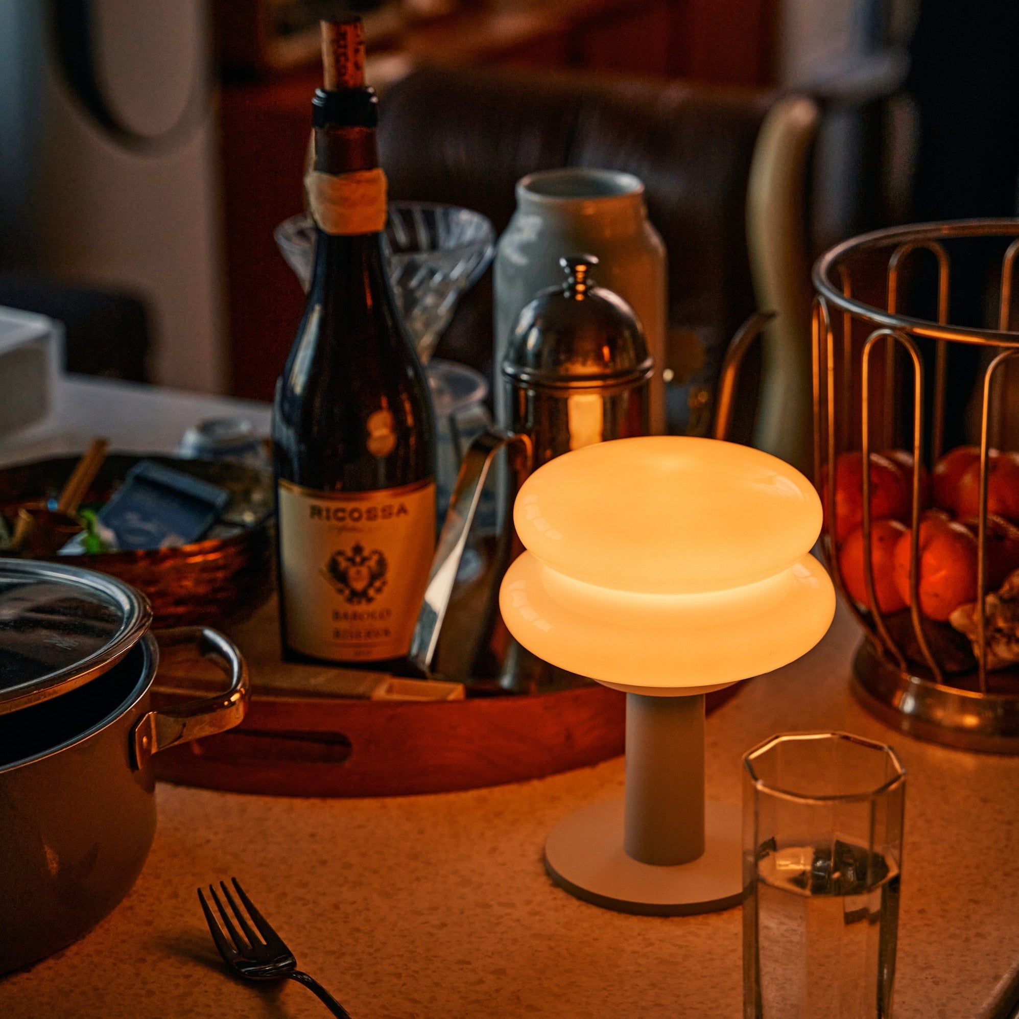 Table setting with a bottle of wine, glass,, portable table lamp and other items on a table.
