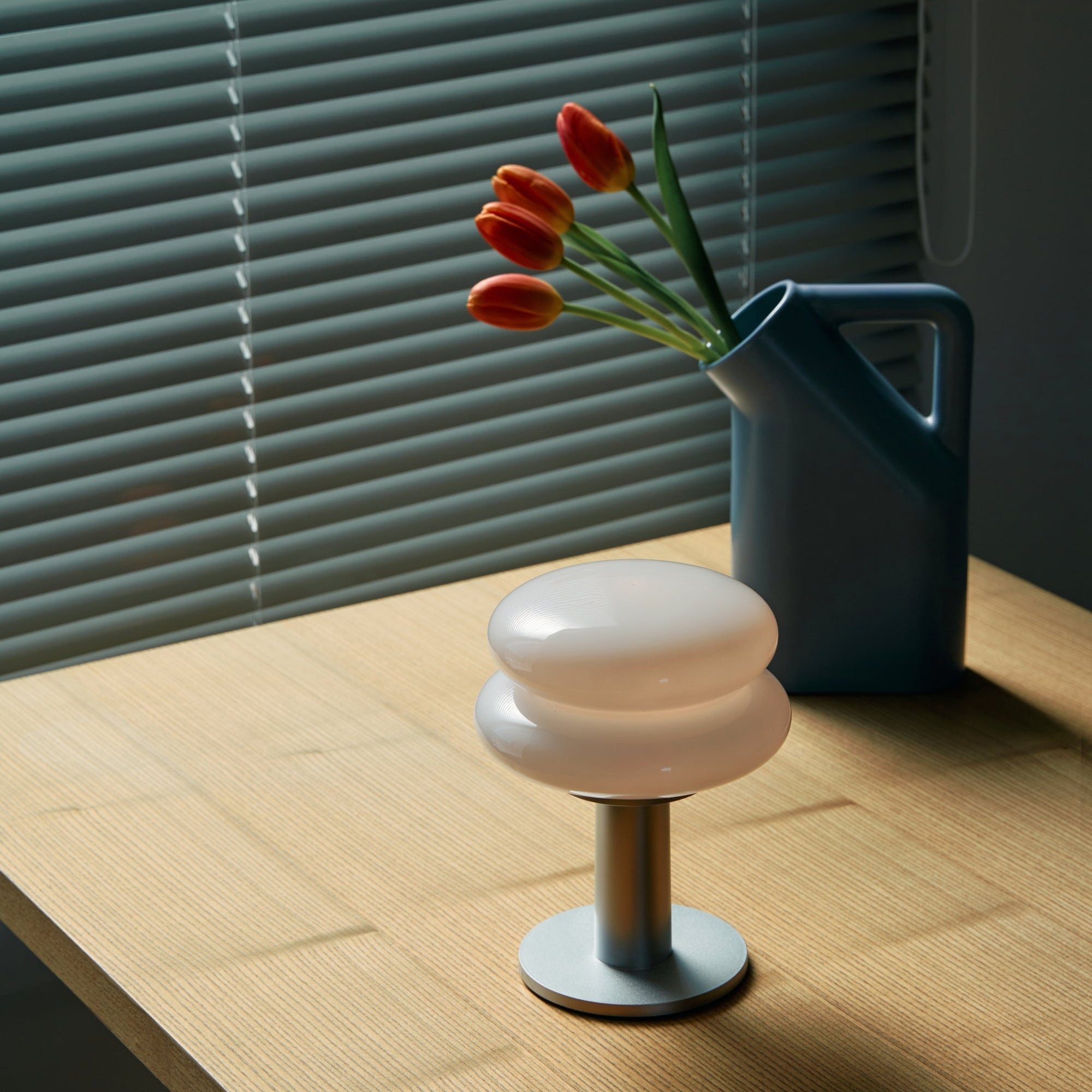 Table with a vase with flowers and a portable table lamp with venetian blinds in the background.