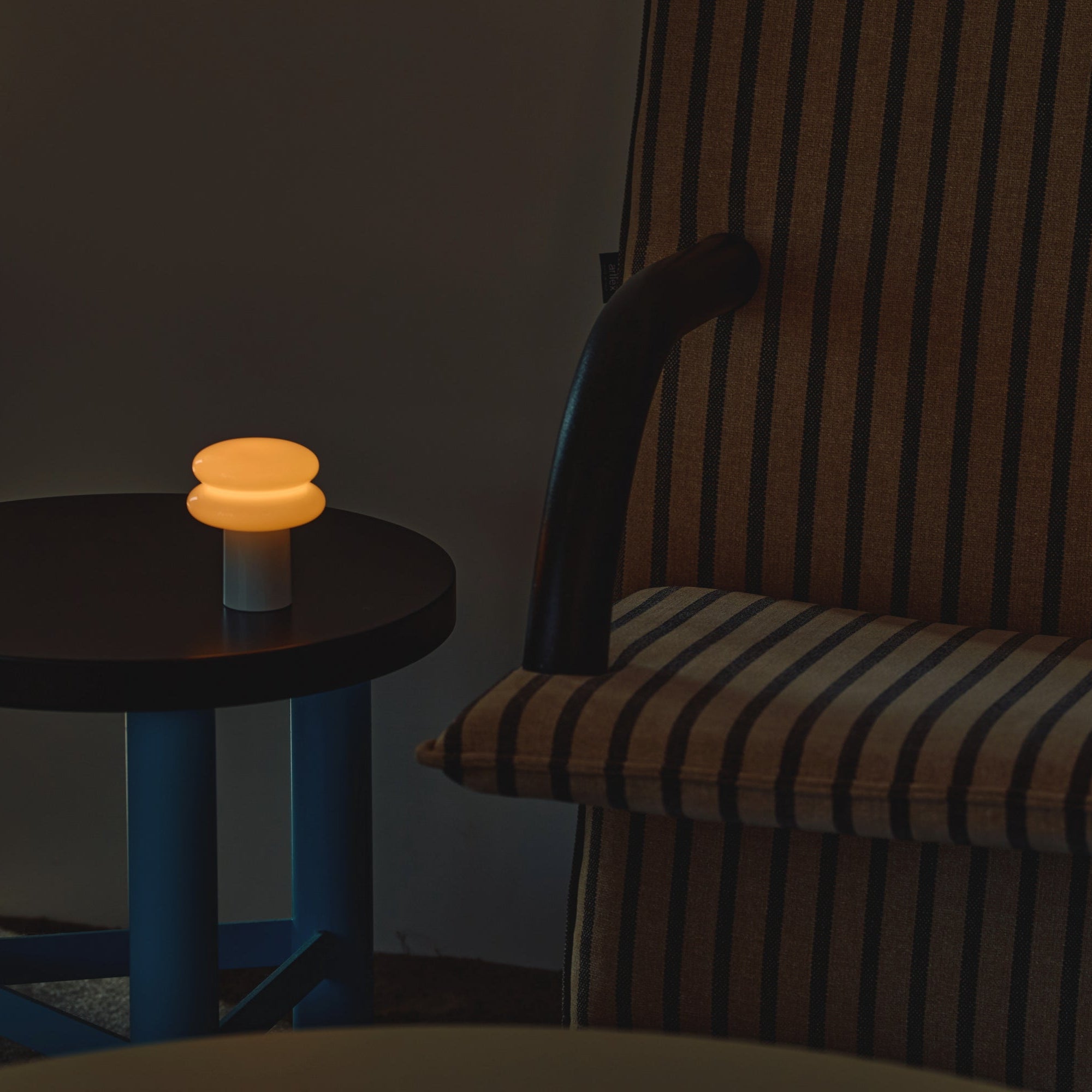 Small round table with a glowing lamp next to a striped armchair in a dimly lit room.