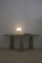 A modern table lamp with a glowing white mushroom-shaped shade and a matching base, placed on a minimalist gray console table against a plain light gray wall.