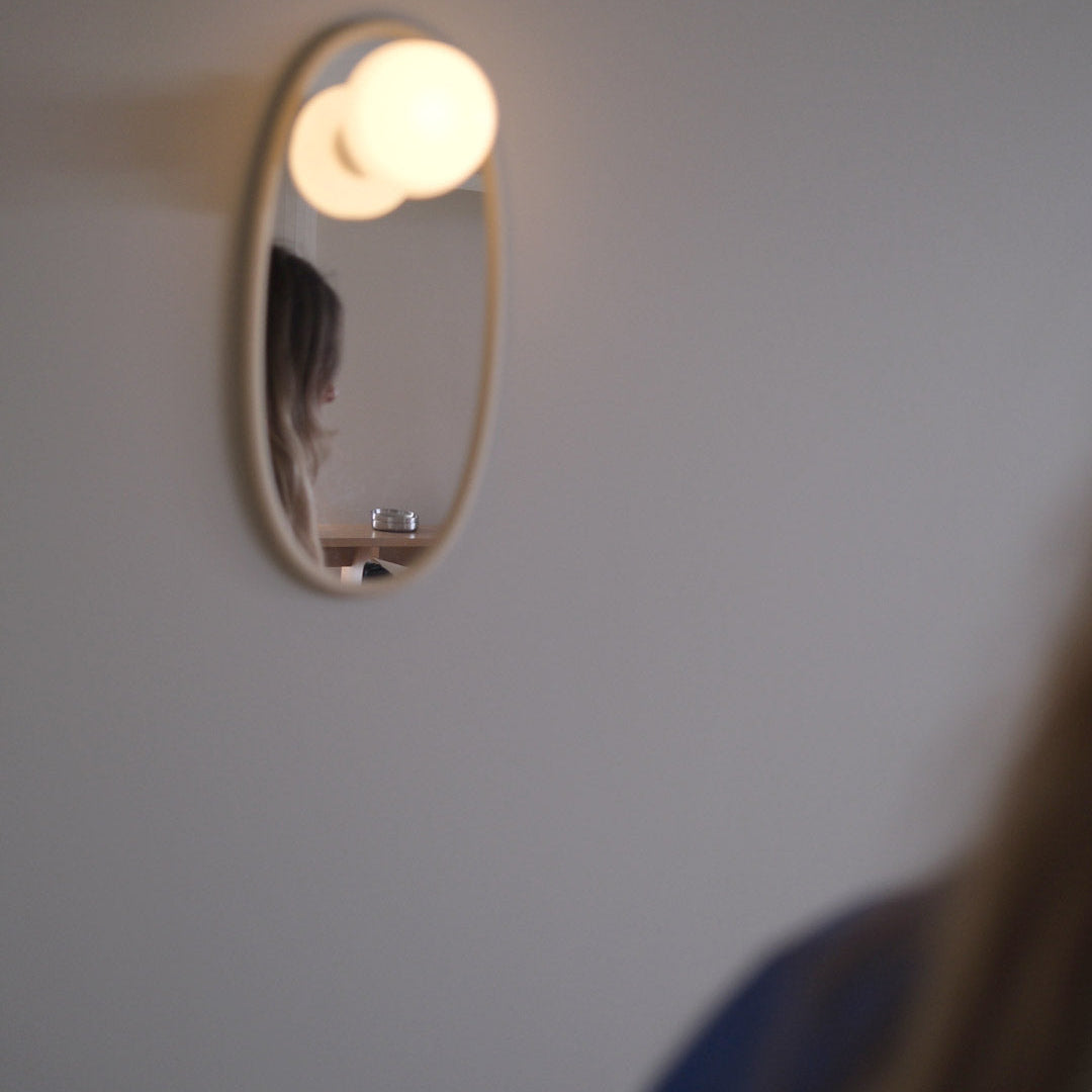 A small oval wall mirror with a warm glowing bulb light above it, mounted on a plain wall. The reflection shows part of a person, and the foreground includes blurred hair and clothing.
