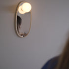 A small oval wall mirror with a warm glowing bulb light above it, mounted on a plain wall. The reflection shows part of a person, and the foreground includes blurred hair and clothing.