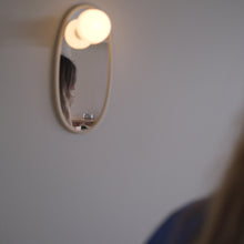 A small oval wall mirror with a warm glowing bulb light above it, mounted on a plain wall. The reflection shows part of a person, and the foreground includes blurred hair and clothing.