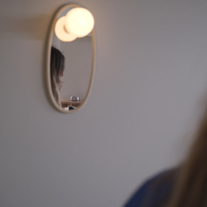 A small oval wall mirror with a warm glowing bulb light above it, mounted on a plain wall. The reflection shows part of a person, and the foreground includes blurred hair and clothing.