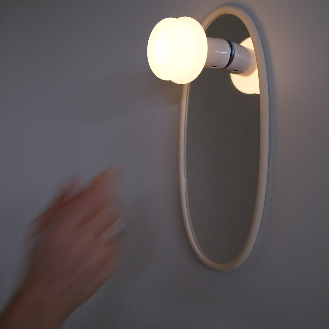 A wall-mounted oval mirror with a glowing frosted bulb attached to its right side, photographed against a plain gray wall. A blurred human hand enters from the left side of the frame.