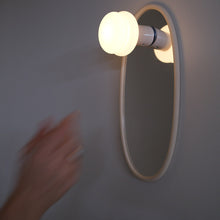 A wall-mounted oval mirror with a glowing frosted bulb attached to its right side, photographed against a plain gray wall. A blurred human hand enters from the left side of the frame.