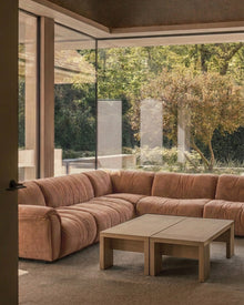 A large L-shaped sectional sofa upholstered in a dusty reddish brown velvet type fabric, positioned in a modern living room with a light wood coffee table and large glass windows overlooking a garden.
