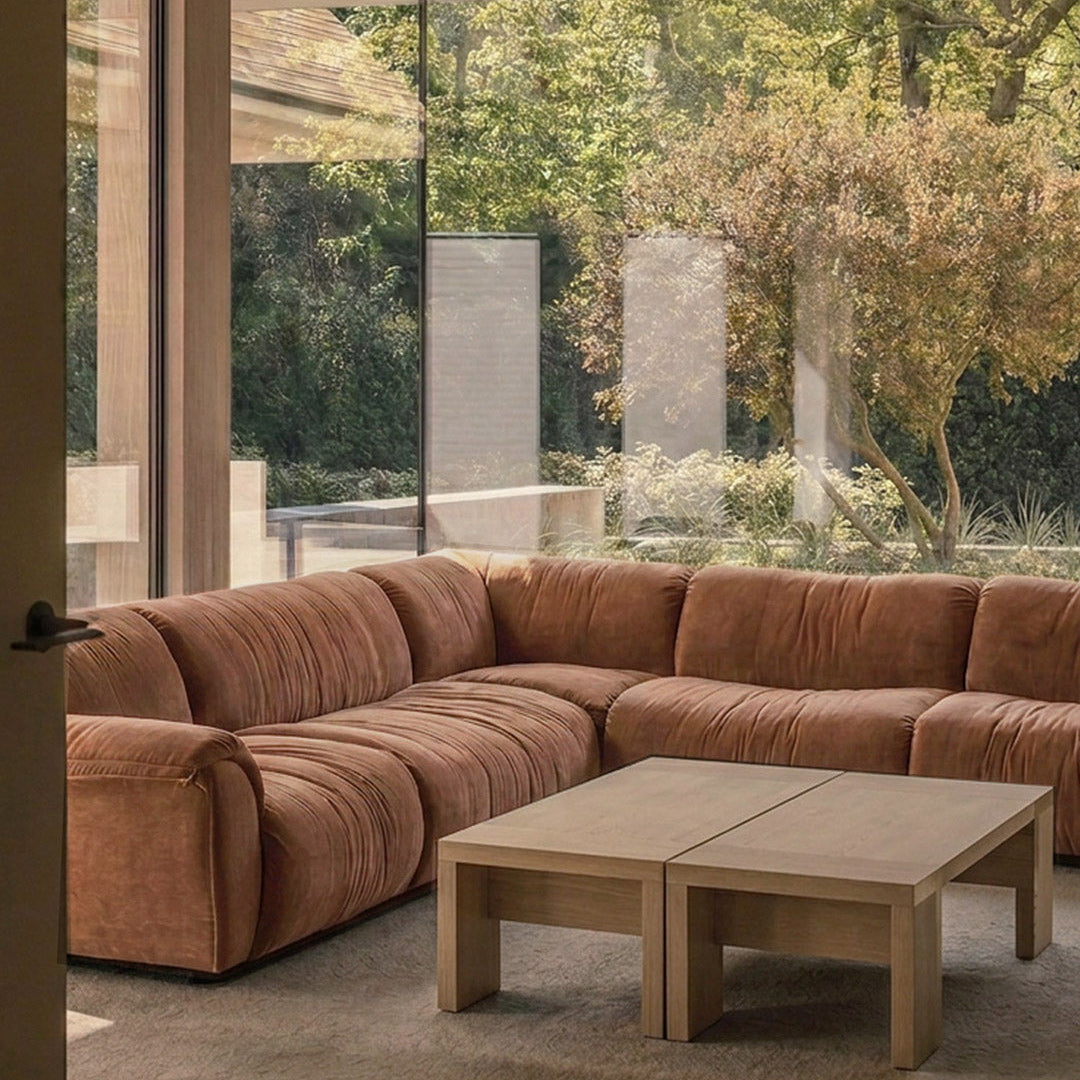 A modern sectional sofa in a warm rust-brown fabric, arranged in an L-shape in a minimalist living room with a light wood coffee table and large glass windows overlooking a garden.