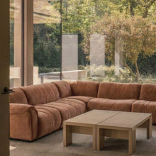 A modern sectional sofa in a warm rust-brown fabric, arranged in an L-shape in a minimalist living room with a light wood coffee table and large glass windows overlooking a garden.
