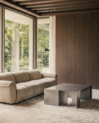 A modern living room featuring a beige upholstered sofa, a low rectangular metallic coffee table, a textured rug, and a wood-paneled wall with large windows overlooking a forest.