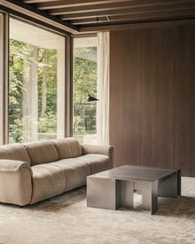 A modern living room featuring a beige upholstered sofa, a low rectangular metallic coffee table, a textured rug, and a wood-paneled wall with large windows overlooking a forest.