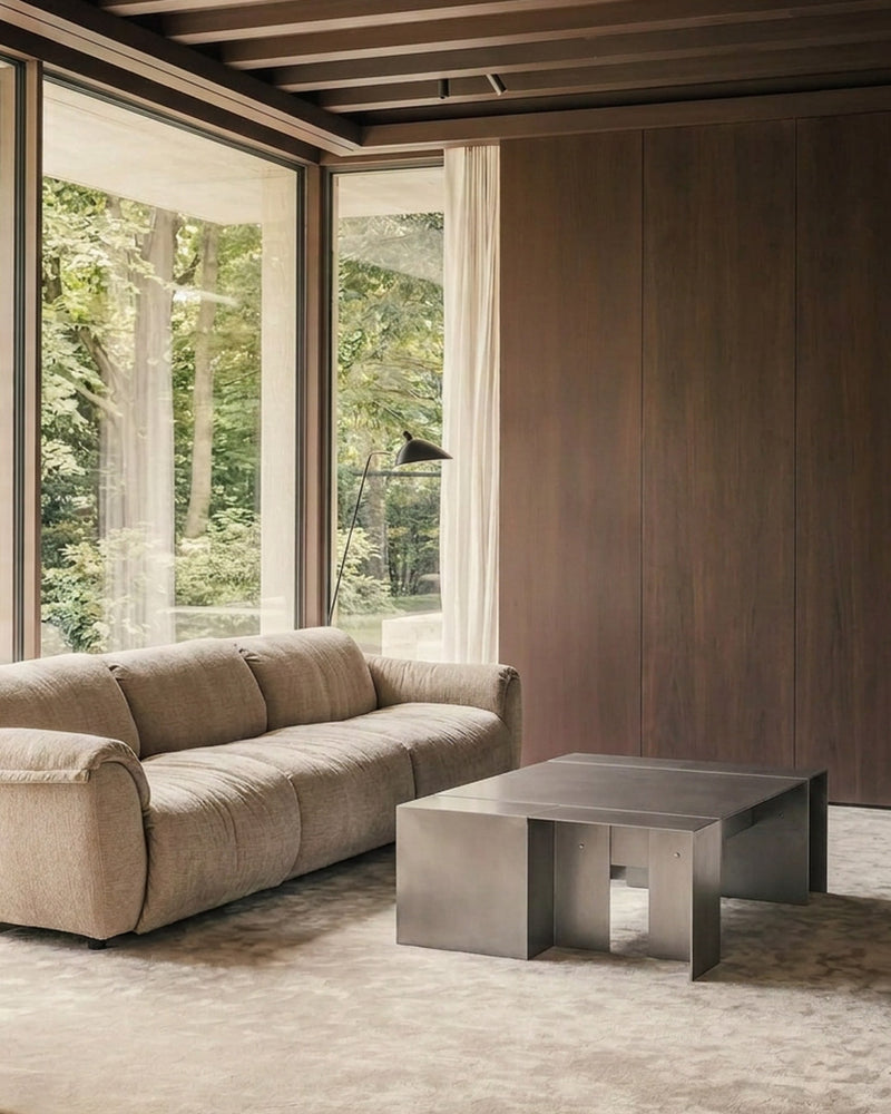 A modern living room featuring a beige upholstered sofa, a low rectangular metallic coffee table, a textured rug, and a wood-paneled wall with large windows overlooking a forest.