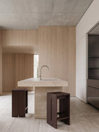 A modern kitchen interior with a light stone island sink, a curved metal faucet, two dark wood stools, and light wood cabinetry in a minimalist architectural space.
