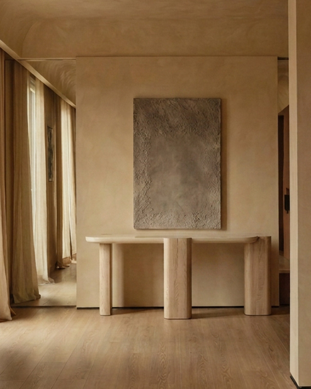 A minimalist interior scene featuring a light wood console table against a textured beige plaster wall, with a large rectangular abstract gray painting mounted above it. Warm wood flooring and draped curtains are visible on the left.