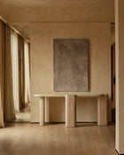 A minimalist interior scene featuring a light wood console table against a textured beige plaster wall, with a large rectangular abstract gray painting mounted above it. Warm wood flooring and draped curtains are visible on the left.