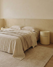 A minimalist beige bedroom scene featuring a neatly made bed with textured linen bedding, a matching beige headboard, a cylindrical side table, and a large plush rug on a light wood floor.