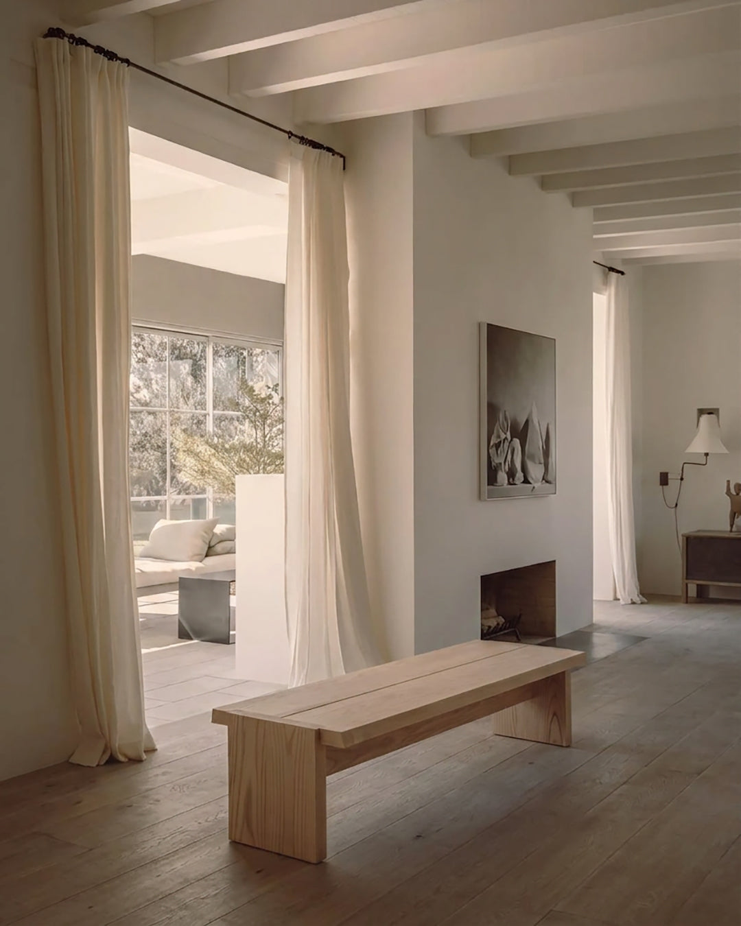 A minimalist light wood bench in a bright, modern room with high ceilings, white walls, and floor-to-ceiling curtains.