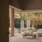 A modern interior seating area with two cream tufted armchairs facing a light wood coffee table, set in a spacious room with textured walls and large glass doors opening to a garden.