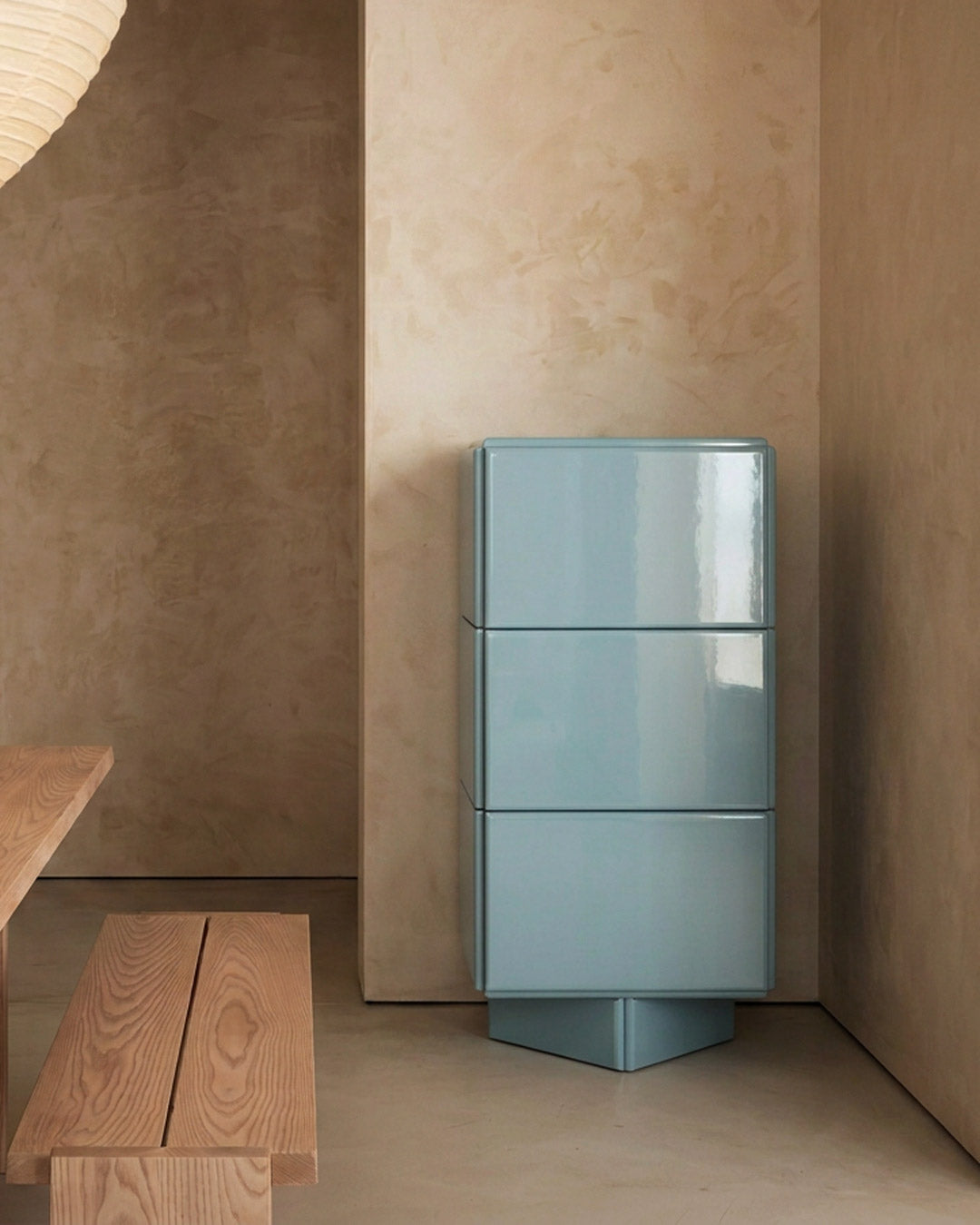 A glossy light blue three-drawer storage cabinet with a geometric base, set against textured beige plaster walls in a minimalist interior.