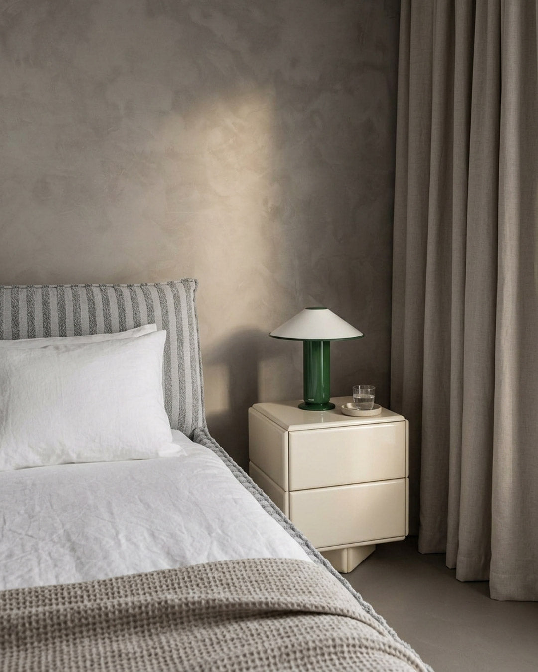 A modern bedroom scene featuring a cream-colored two-drawer nightstand with a green glass table lamp, set next to a bed with white linens and a textured gray headboard against a textured plaster wall.