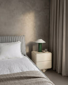 A modern bedroom scene featuring a cream-colored two-drawer nightstand with a green glass table lamp, set next to a bed with white linens and a textured gray headboard against a textured plaster wall.