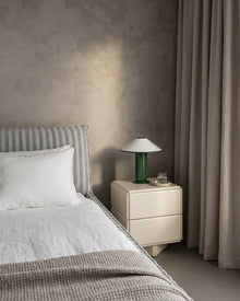 A modern bedroom scene featuring a cream-colored two-drawer nightstand with a green glass table lamp, set next to a bed with white linens and a textured gray headboard against a textured plaster wall.