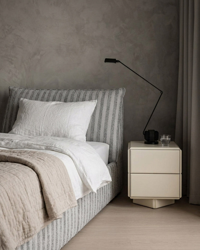 A modern bedroom scene featuring a bed with a gray and white vertically striped upholstered headboard and matching footboard, dressed in white linens and a beige quilt. A cream-colored two-drawer nightstand with a black adjustable floor lamp stands beside it against a textured gray plaster wall.